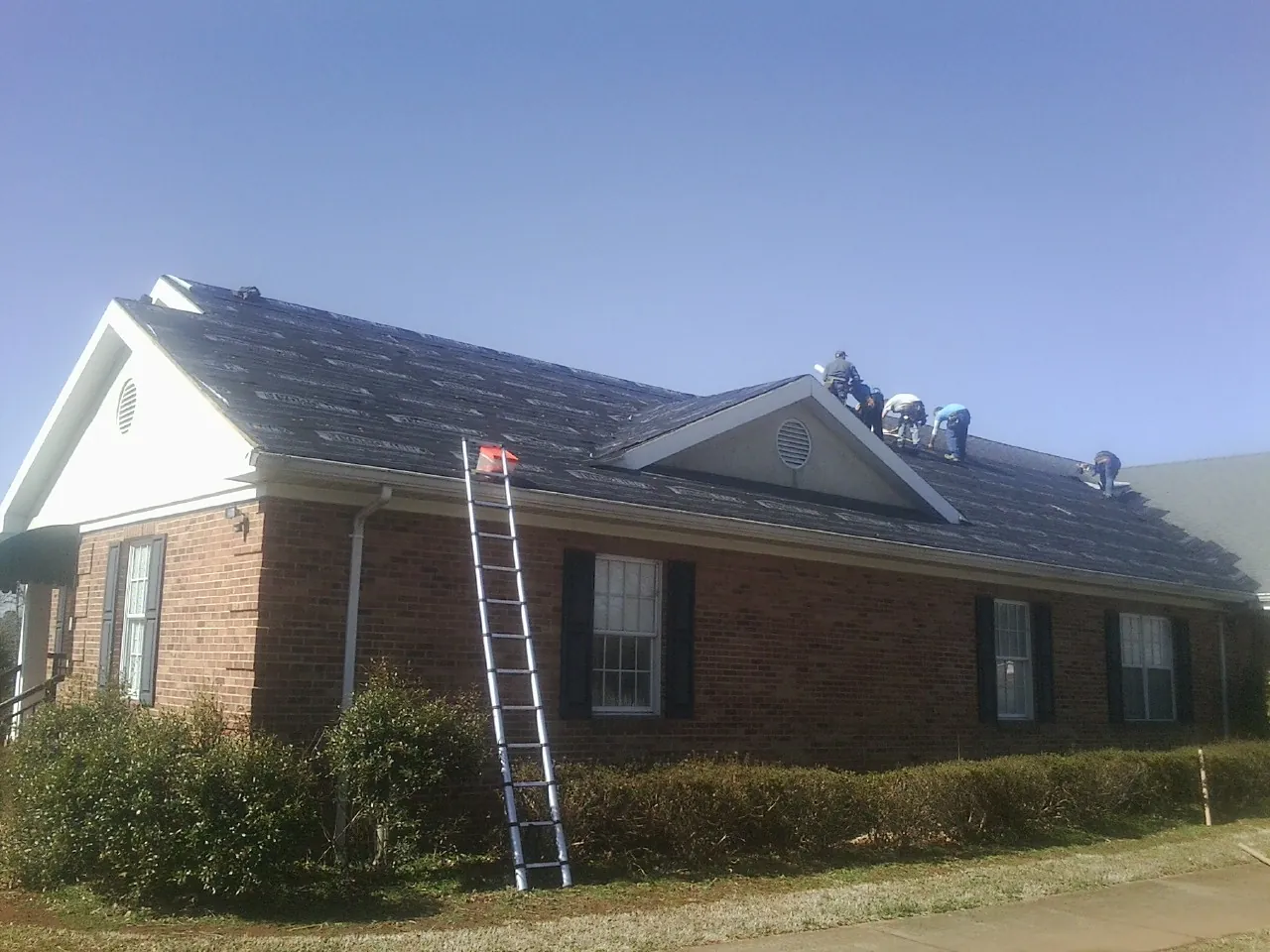 Crew tearing shingles off a brick ranch roof with a ladder set — Metal Roof Repair work by our crew in Fork