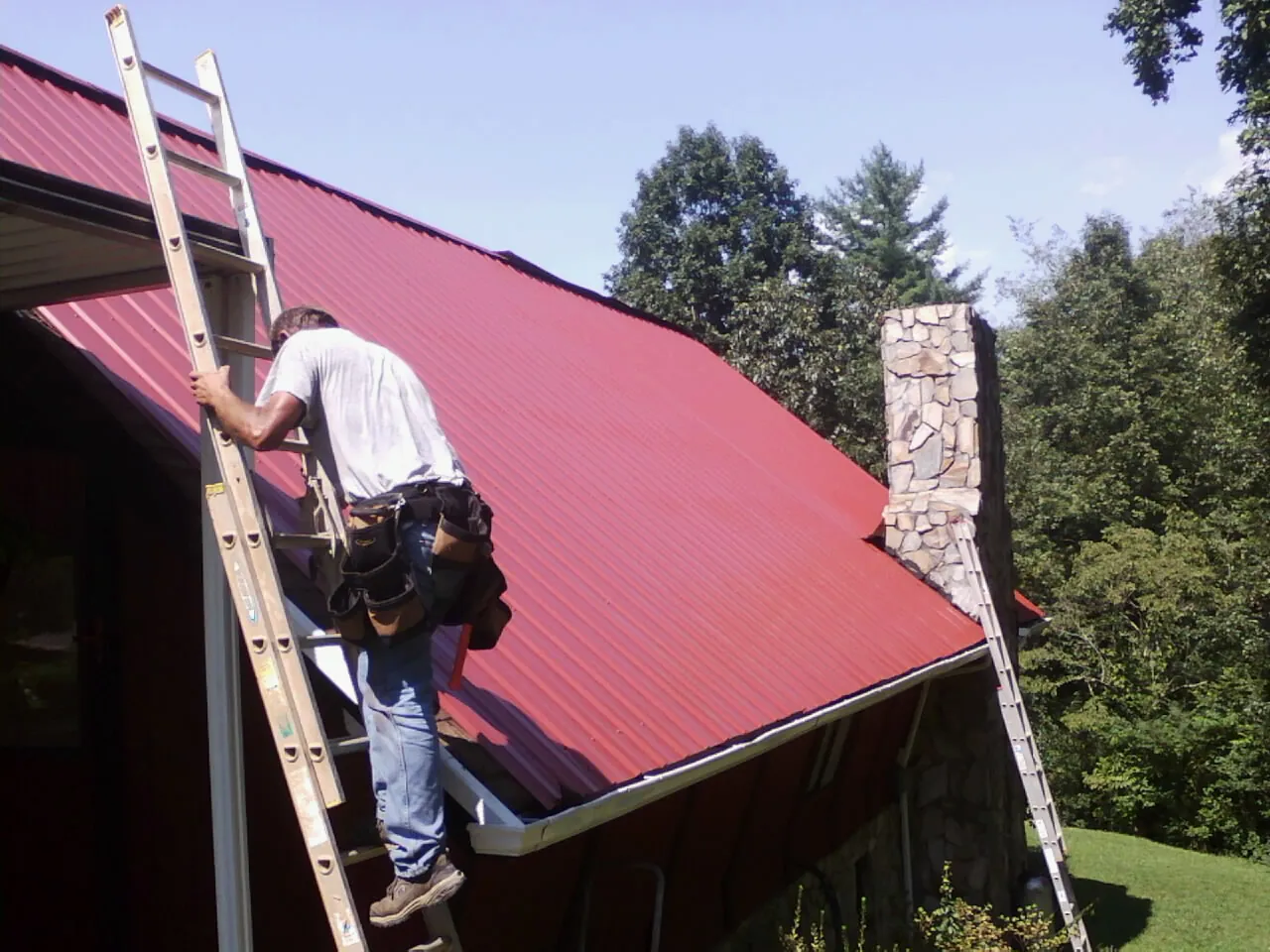 Roofer on a ladder beside a red metal roof and stone chimney — Soffit & Fascia Repair by Roofing Brothers in Fork